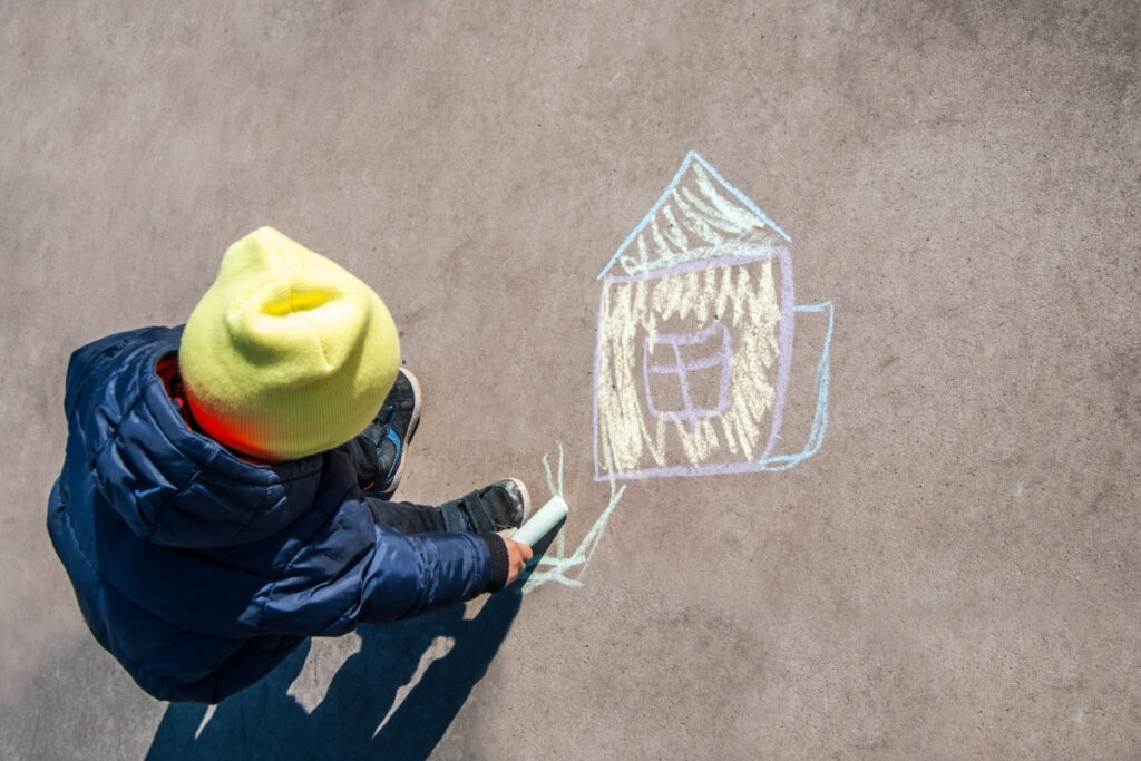 A child draws a chalk picture of a house on the pavement.