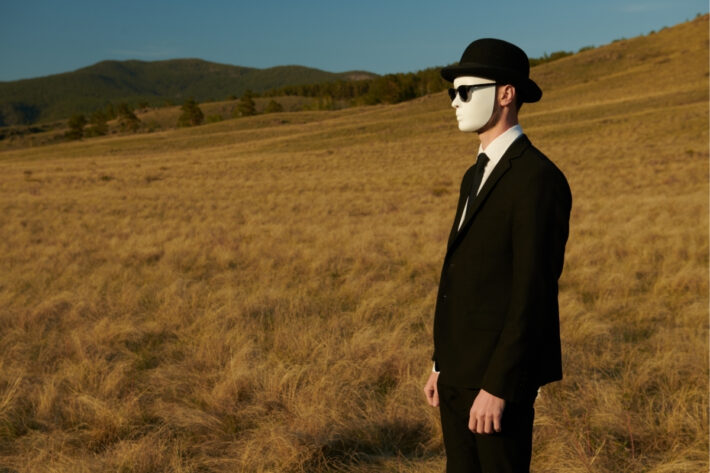 A masked man wearing a suit, hat, and sunglasses stands in a desolate field.